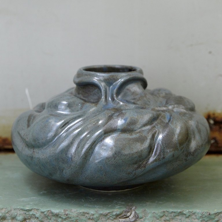 Christina Muff, dansk samtidskeramiker (f. 1971). Skulpturel vase dekoreret med 
spirende vækster. Denne unikavase er glaseret med
silkemat stålblå glasur der bryder i brunlige toner.